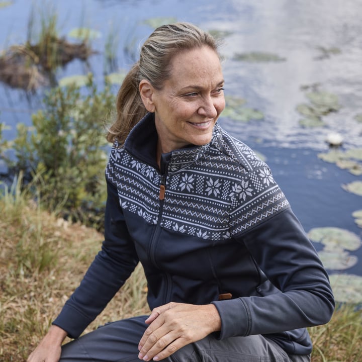 Eine Frau mit hellbraunem Haar, das zu einem Pferdeschwanz gebunden ist, sitzt neben einem Teich, der mit Seerosenblättern übersät ist, trägt eine dunkle Jacke mit weißem Nordic-Muster und schenkt ein sanftes Lächeln, während sie zur Seite blickt.