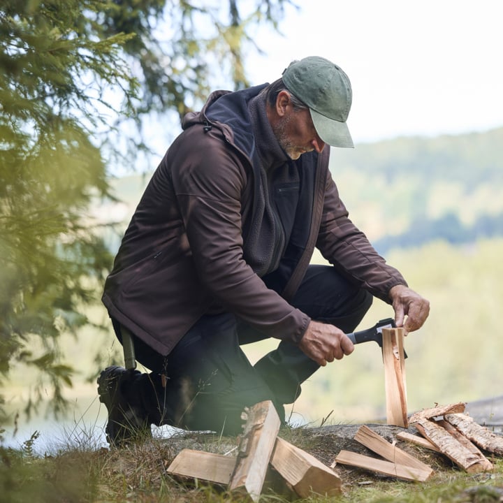 Ein Mann mit grüner Mütze und brauner Jacke kniet draußen und hackt Feuerholz mit einer Axt. Um ihn herum liegen gespaltene Baumstämme, mit Bäumen und einem bewaldeten Gebiet im Hintergrund.