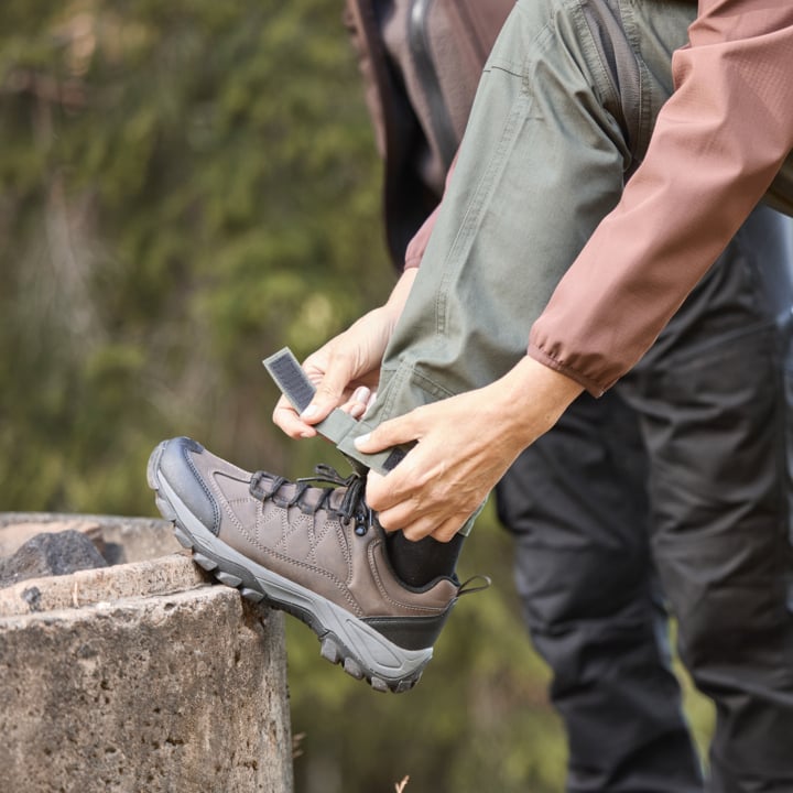 In Outdoor-Kleidung zieht man den Riemen am Bündnis der grünen Hose fester und legt einen Stiefel auf Beton. Der Hintergrund zeigt verschwommene Bäume.