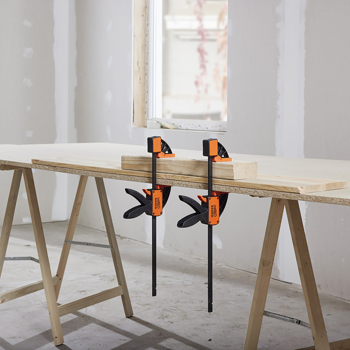 Ein Paar orange-schwarze Klemmen halten ein Holzbrett an einem größeren Stück Holz, beide ruhen auf zwei Sägeböcken in einem teilweise fertigen Raum mit freiliegender Trockenbauwand und einem Fenster.