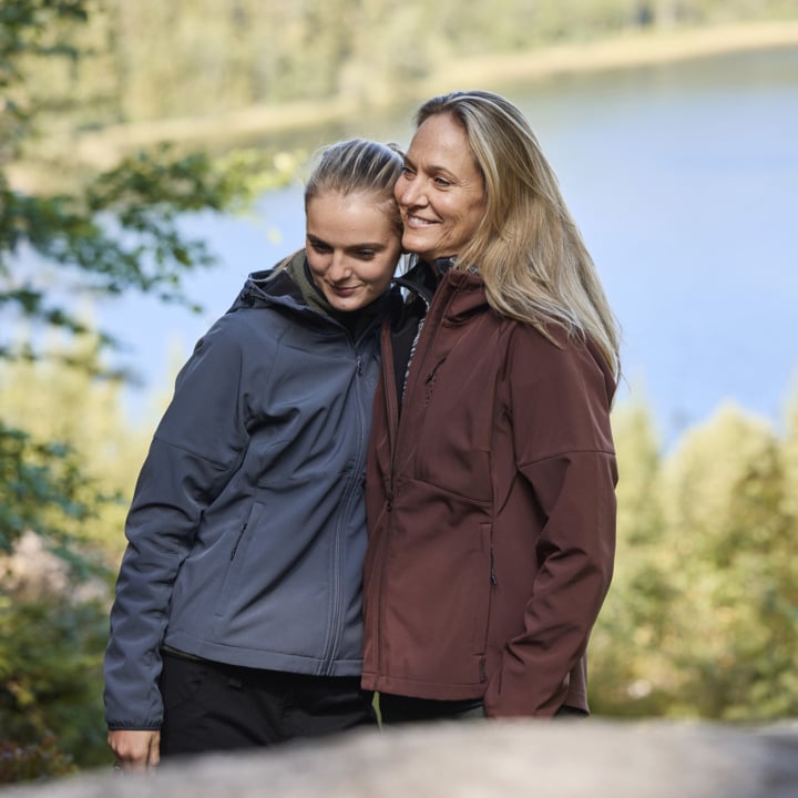 To kvinner, en i grå jakke og den andre i burgunder, står tett sammen og smiler. De står utenfor ved en innsjø omgitt av trær og frodig grønt.