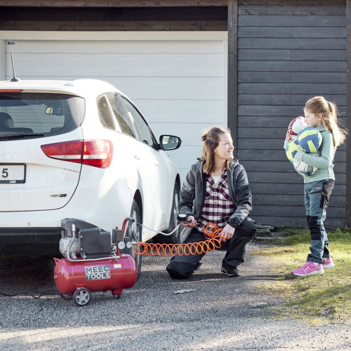 Eine Frau kniet neben einem weißen Auto und pumpt einen Reifen mit einem Luftkompressor auf. Ein junges Mädchen steht in der Nähe und hält Sportbälle. Sie stehen draußen neben einer Holzgarage.