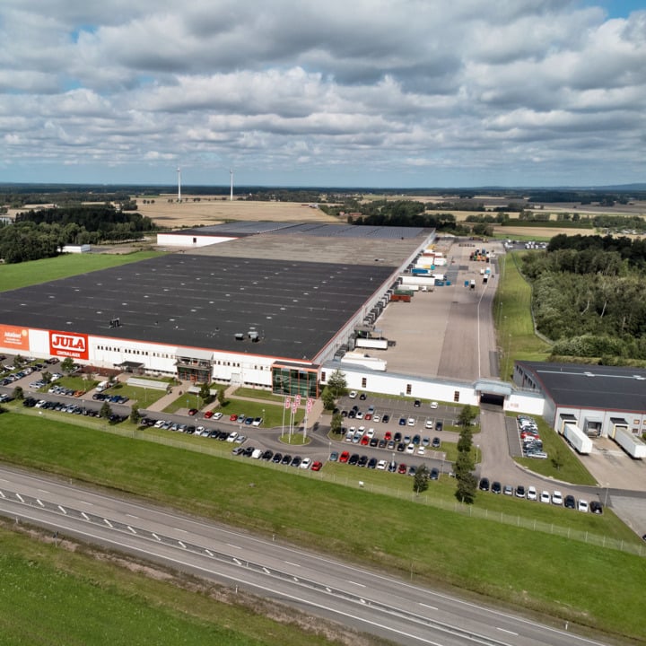 Ett stort lagerhus med en skylt där det står "Jula" på framsidan, omgivet av parkerade bilar och lastbilar. Byggnaden ligger intill en motorväg, med öppna fält, träd och vindkraftverk synliga i fjärran under en delvis molnig himmel.