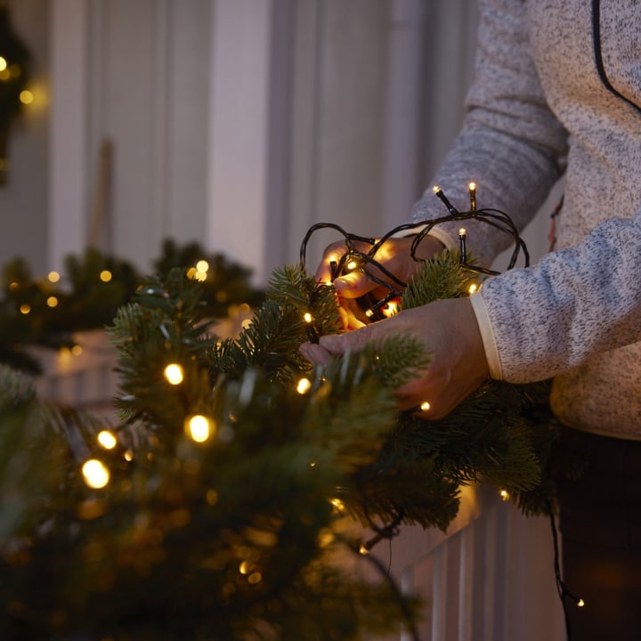 Eine Person, die warme weiße Lichterketten um eine grüne, künstliche Weihnachtsgirlande wickelt.